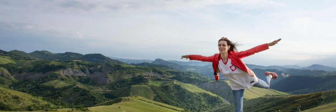 A woman joyfully balances on a rock amidst rolling green hills under a clear blue sky.