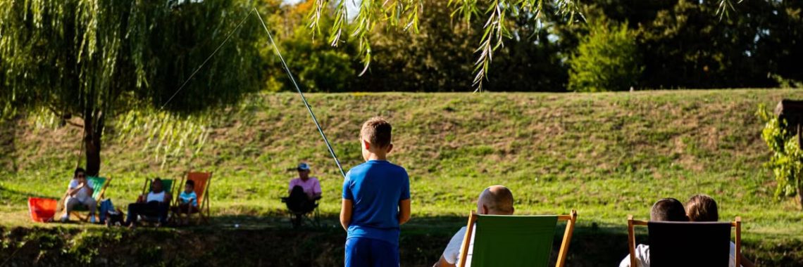 Young boy fishing by a pond on a sunny day, surrounded by trees and relaxed people.