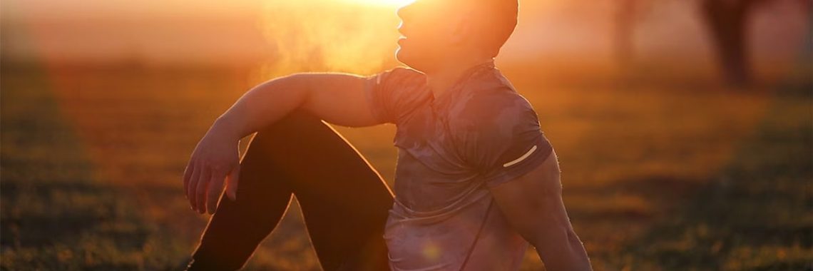 Muscular man sits in rural field at sunset, exhaling visible breath.